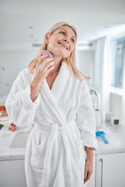 Beautiful adult woman holding skin care device while doing\
massage on neck in room indoors