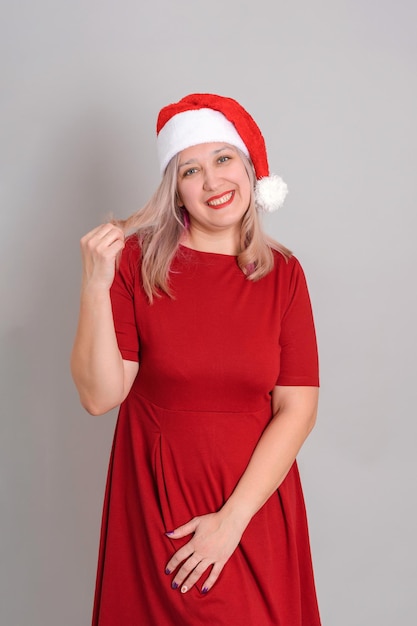 A beautiful adult woman in a Santa hat and a red dress poses on a gray background