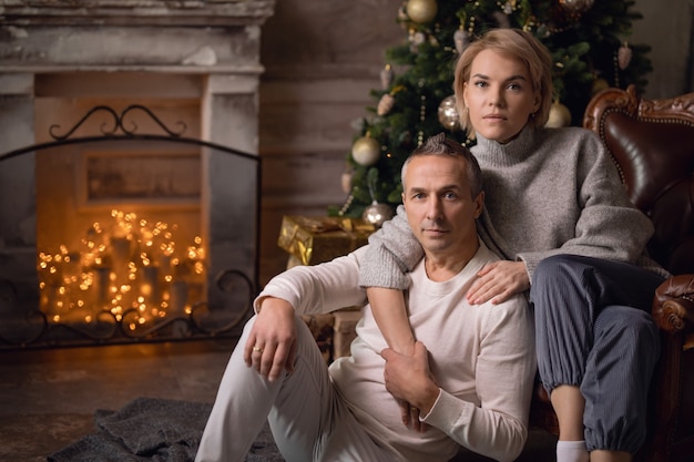 beautiful adult woman with a man sit on an armchair and hug near a christmas tree and fireplace