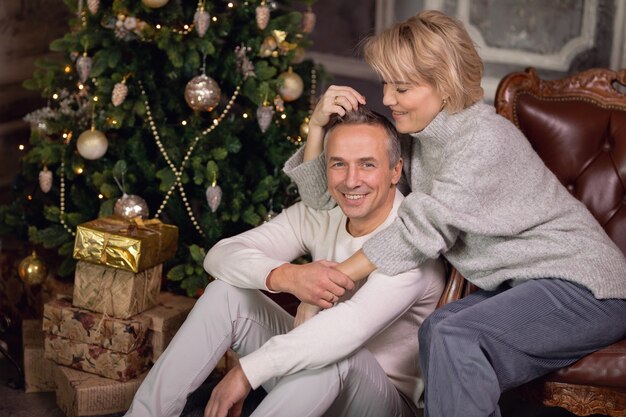 beautiful adult woman with a man sit on an armchair and hug near a christmas tree