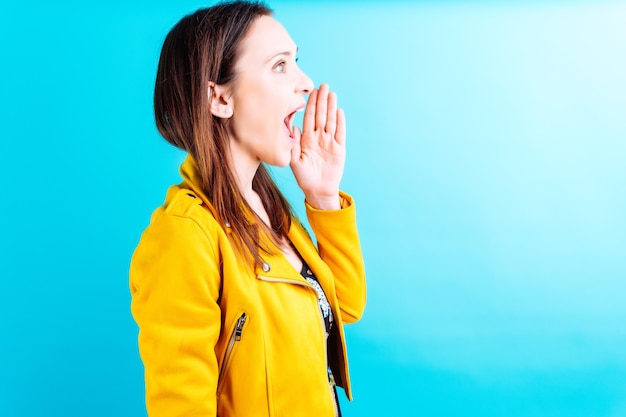 Beautiful adult young woman in butterfly summer dress and\
yellow jacket on blue background talking loudly with hand on mouth.\
concept speak loud or shout