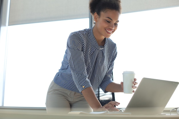 Красивая афро-американская бизнес-леди в умной повседневной одежде работает на ноутбуке в офисе.