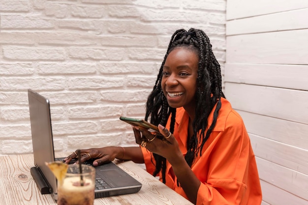 スマートフォンで通話しながらノートパソコンを操作する美しいアフリカ系アメリカ人女性のフリーランサー リモートでの作業のコンセプト
