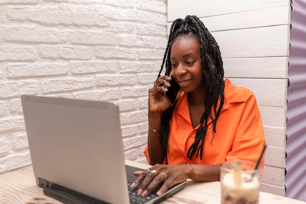 Beautiful african american female freelancer working with laptop while talking on a call with smartphone Concept of working on remote