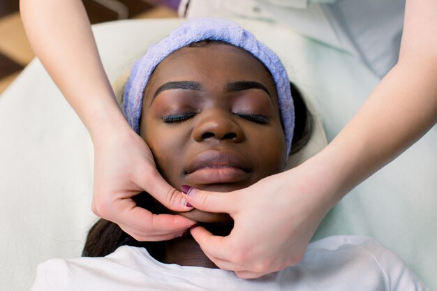 Beautiful African-American girl having facial massage with closed eyes in spa salon