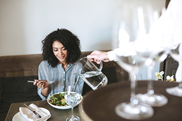 Beautiful African American girl sitting in restaurant and eating salad while waiter pouring water in glass