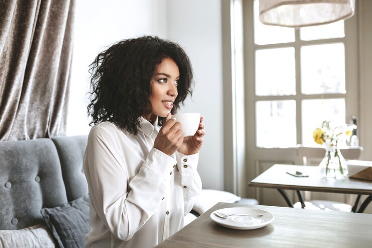 Beautiful African American girl sitting in restaurant with cup in hands.Young pretty lady in white shirt drinking coffee in cafe