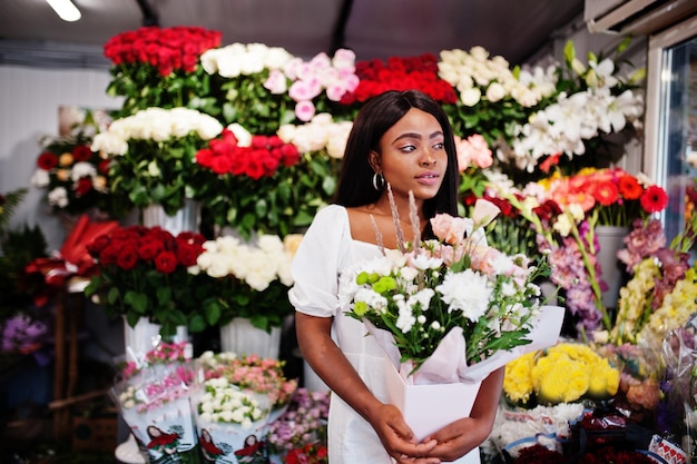 Beautiful african american girl in tender white dress with bouquet flowers in hands standing against floral background in flower shop.Black female florist.