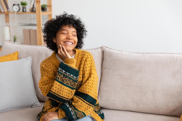 Beautiful african american girl with afro hairstyle smiling sitting on sofa at home indoor Young african woman with curly hair laughing Freedom happiness carefree happy people concept