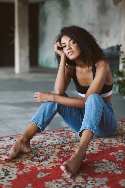 Beautiful african american girl with dark curly hair in black sporty top and jeans thoughtfully  while sitting on vintage carpet at home