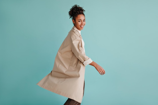 Beautiful african american lady in trench coat looking at camera Studio shot of blithesome black young woman posing on turquoise background