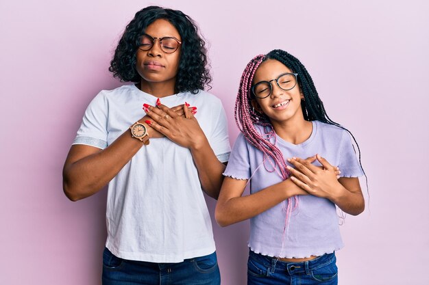 Beautiful african american mother and daughter wearing casual clothes and glasses smiling with hands on chest with closed eyes and grateful gesture on face health concept