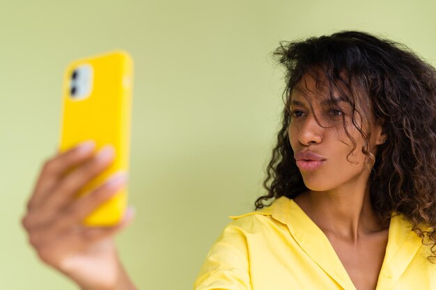 緑の背景にカジュアルなシャツを着た美しいアフリカ系アメリカ人女性が、スマートフォンで自撮り写真を撮って幸せです