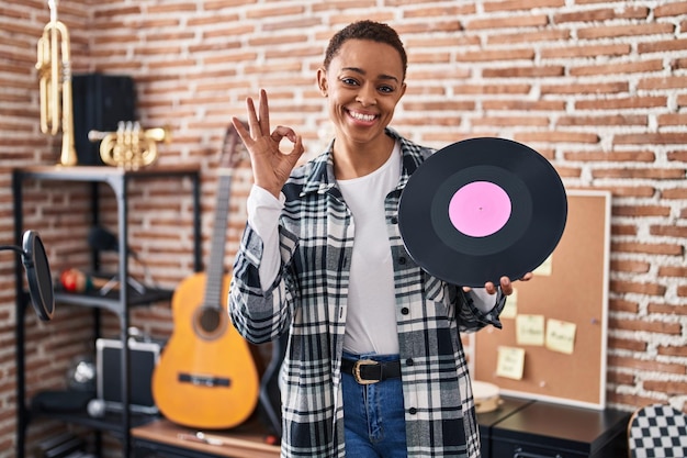 音楽スタジオでビニール レコードを保持している美しいアフリカ系アメリカ人女性が指で ok サインをし、フレンドリーなジェスチャーの優れたシンボルに笑みを浮かべて