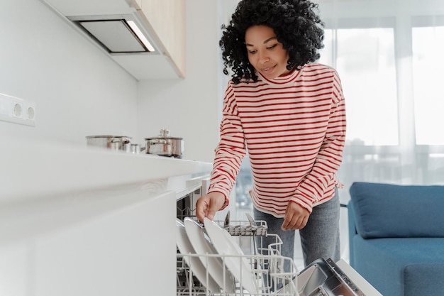 beautiful african american woman wearing casual clothes loading dishwasher with dishes in kitchen