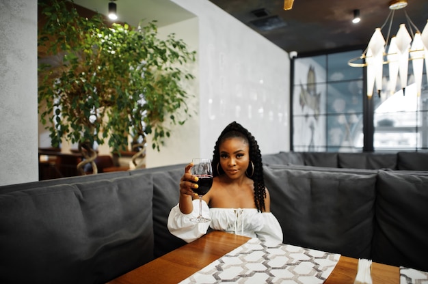 Beautiful african american woman in white blouse and red leather pants pose at restaurant with glass of wine.