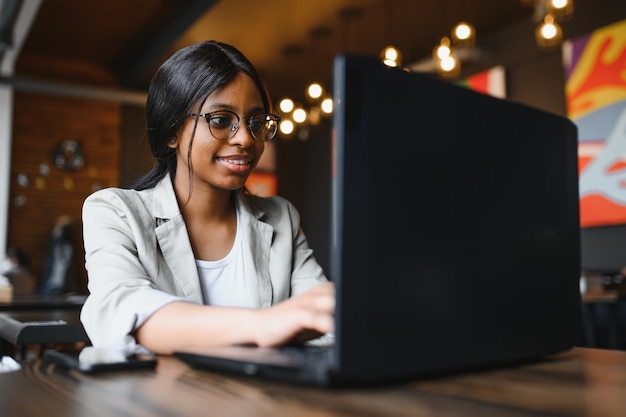 Красивая африканская деловая женщина ждет встречи в коворкинге и сидит с портативным компьютером Девушка-хипстер просматривает веб-страницы на ноутбуке во время обеда