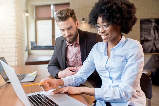 Beautiful african businesswoman and caucasian man working together with laptops near the window at the cafe or office