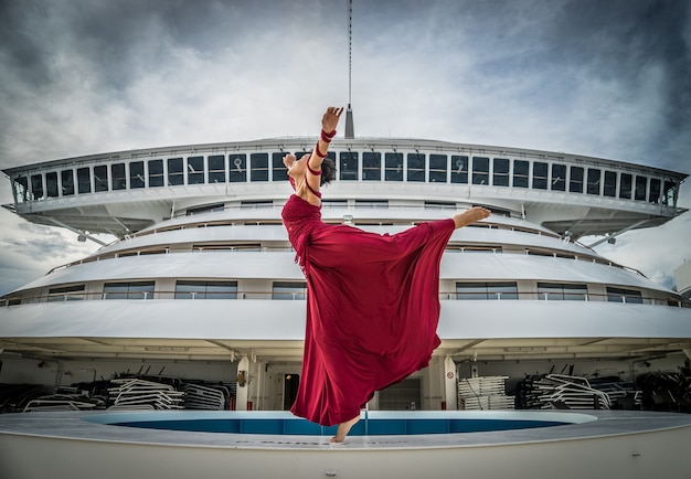 Beautiful african dancer with black hair wearing red dress dancing on the bow of the ship on the bridge on background.