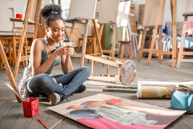 Beautiful african ethnicity student sitting with coffee on the floor with still life painting at the university classroom