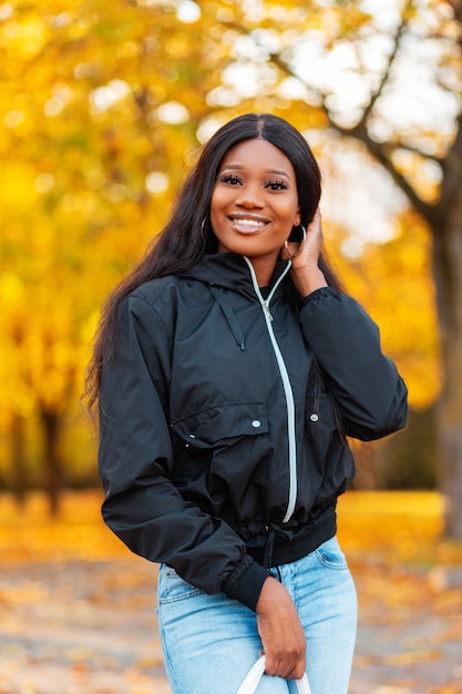 Bella ragazza africana in giacca e jeans che cammina nel parco con foglie autunnali dorate