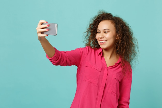 Bella ragazza africana in abiti casual rosa facendo selfie girato sul telefono cellulare isolato su sfondo blu turchese parete in studio. persone sincere emozioni, concetto di stile di vita. mock up copia spazio.