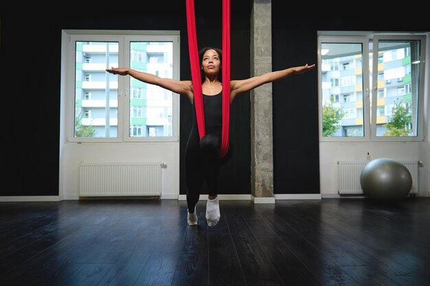 Beautiful African sporty woman hanging on a red hammock with outstretched hands in a fitness and yoga studio. Slim body young woman practicing aerial flying yoga with anti-gravity