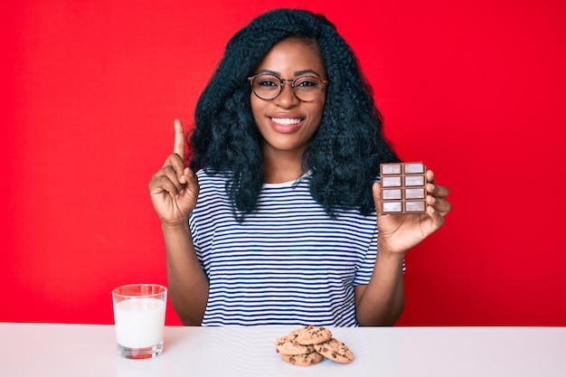 チョコレートバーを食べて、幸せそうな顔で指を指しているアイデアや質問に微笑んで牛乳を飲む美しいアフリカの女性