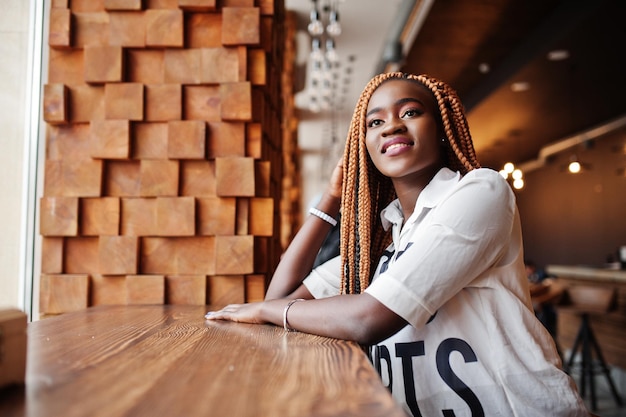 Beautiful african woman in stylish casual shirt and dreadlocks posing at cafe near the window sill