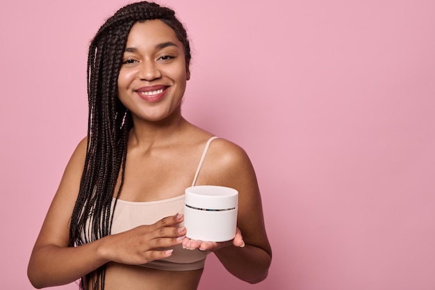 Beautiful African woman with stylish dreadlocks smiles looking at camera, holding a container with moisturizer in her hand