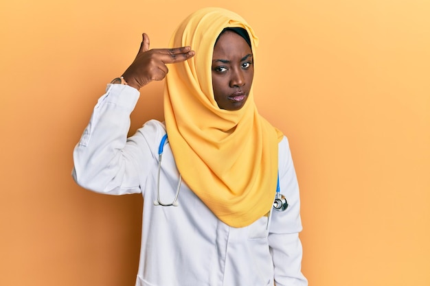 Beautiful african young woman wearing doctor uniform and hijab shooting and killing oneself pointing hand and fingers to head like gun, suicide gesture.