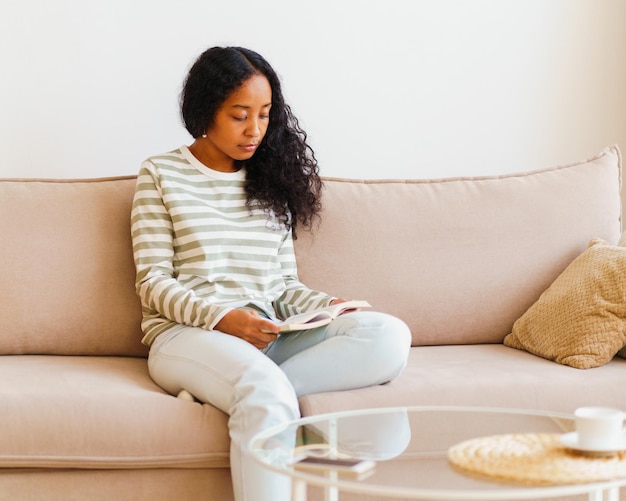 Photo beautiful africanamerican female sitting on couch and reading book spending time at home alone