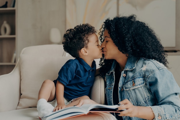 Photo beautiful afro american family mother with little son kissing each other and expressing love while spending time together at home, reading book while sitting on soft white couch in living room