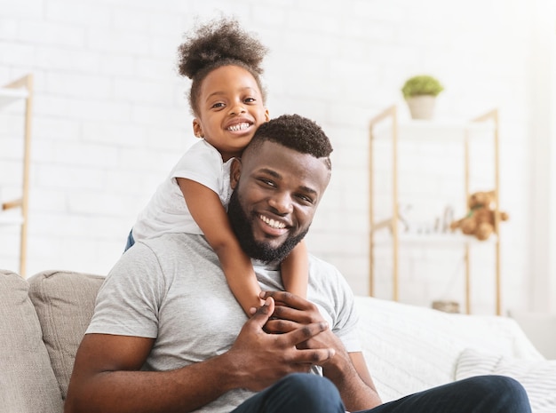 Beautiful afro family of young father and preschool daughter posing at home, afro girl hugging dad from behind, copy space