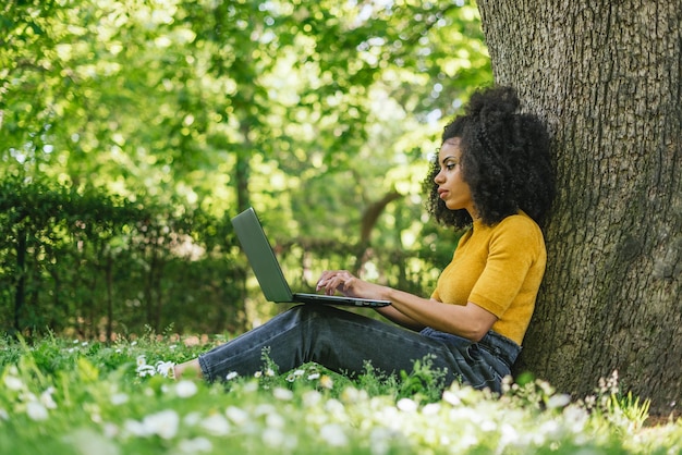 Beautiful afro woman typing on a laptop in a garden. Side view at grass level.