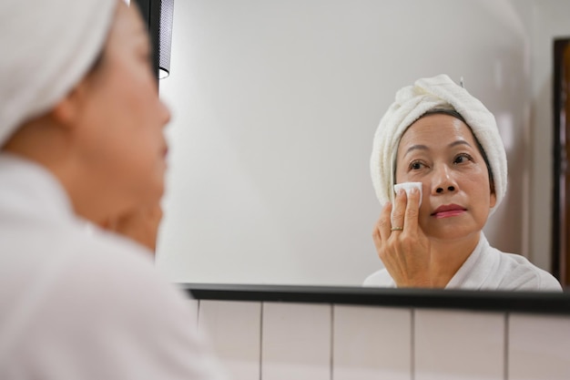 Beautiful aged Asian woman cleansing her face with cotton pad and looking in the mirror