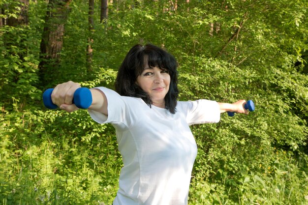 Beautiful aged woman goes in for sports in the park holds dumbbells in her hands and smiles