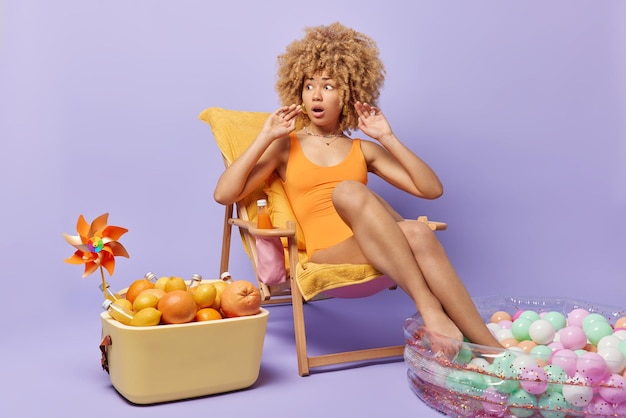 Beautiful amazed woman with curly hair reacts to shocking news\
dresed in swimsuit poses on comfortable deck chair keeps legs in\
inflated pool uses portable fridge isolated over purple\
background