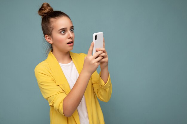 Bella ragazza stupita che indossa giacca gialla e t-shirt bianca in piedi isolato su sfondo blu navigando su internet tramite telefono guardando lo schermo del cellulare.