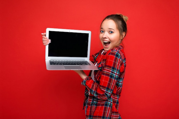 Beautiful amazed young woman holding laptop wearing red shirt looking at camera isolated on red background