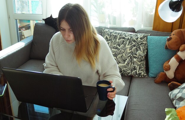 Фото Красивая и серьезная молодая женщина сидит на диване с мягкими игрушками и работает с ноутбуком дома с чашкой кофе концепция удаленной работы дома