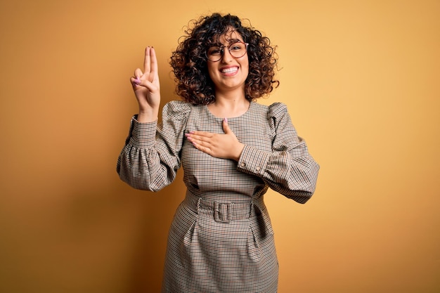 Photo beautiful arab business woman wearing dress and glasses standing over yellow background smiling swearing with hand on chest and fingers up making a loyalty promise oath