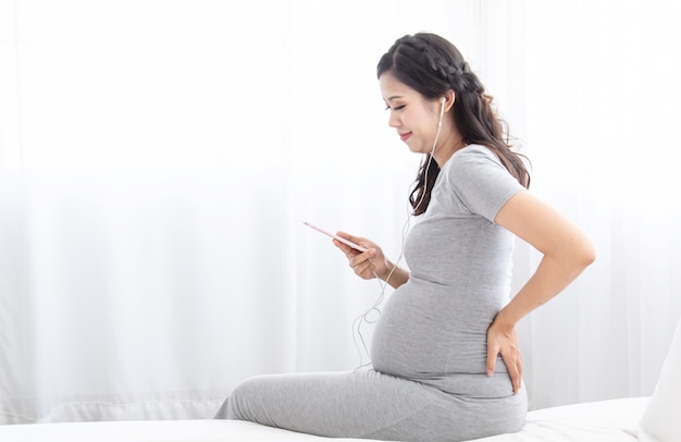 아름 다운 아시아 뱃속 임신 한 여자는 행복 명랑과 헤드셋 또는 헤드폰으로 음악을 듣고, 기대 아가씨는 곧 집에서 침실, 임신 라이프 스타일 개념을 낳을 출산을 줘