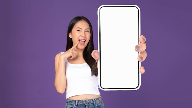 白いタンクトップの美しいアジアのブルネットの女性かわいい女の子空白の画面と白い画面の分離された紫色の背景のモックアップ画像で大きなスマートフォンを示す興奮した驚きの女の子