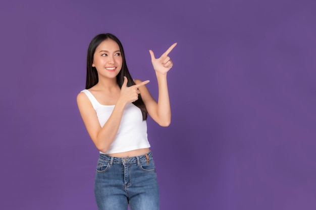 Beautiful Asian brunette woman cute girl in white tank top smiling and pointing finger to empty copy space for present product or blank space for advertising isolated on purple background