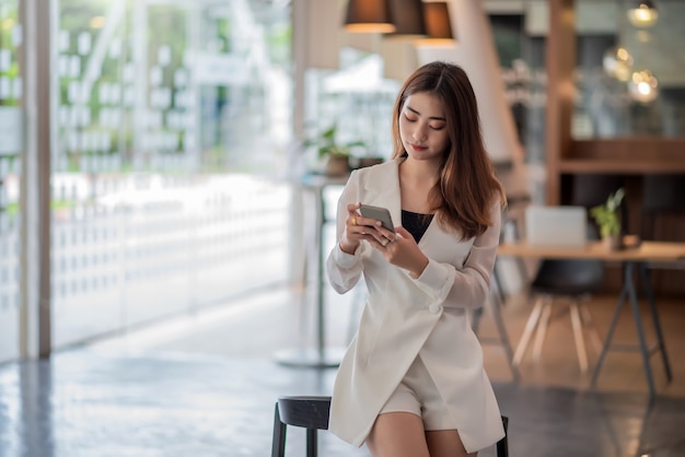 Красивая азиатская бизнес-леди стоя с помощью смартфона в современном офисе.