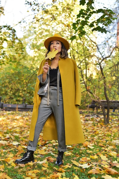 Красивая азиатская деловая женщина в сером костюме, желтой шляпе и желтом пальто в парке рядом с озером Осенний