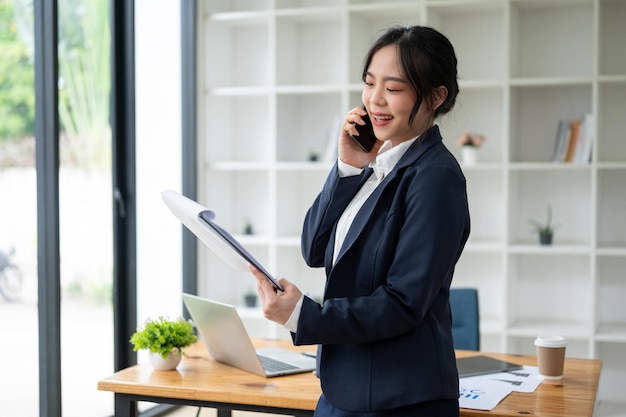 아름다운 아시아 사업가 여성이 전화로 이야기하는 동안 클립보드에서 세부 사항을보고 있습니다.