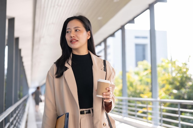 A beautiful Asian businesswoman is walking along a skywalk in the city with her takeaway coffee cup