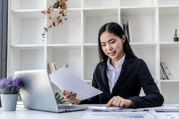 Beautiful Asian businesswoman sitting in her private office, chatting with her partner via laptop and checking documents, she is a female executive of a startup company. Financial management concept.